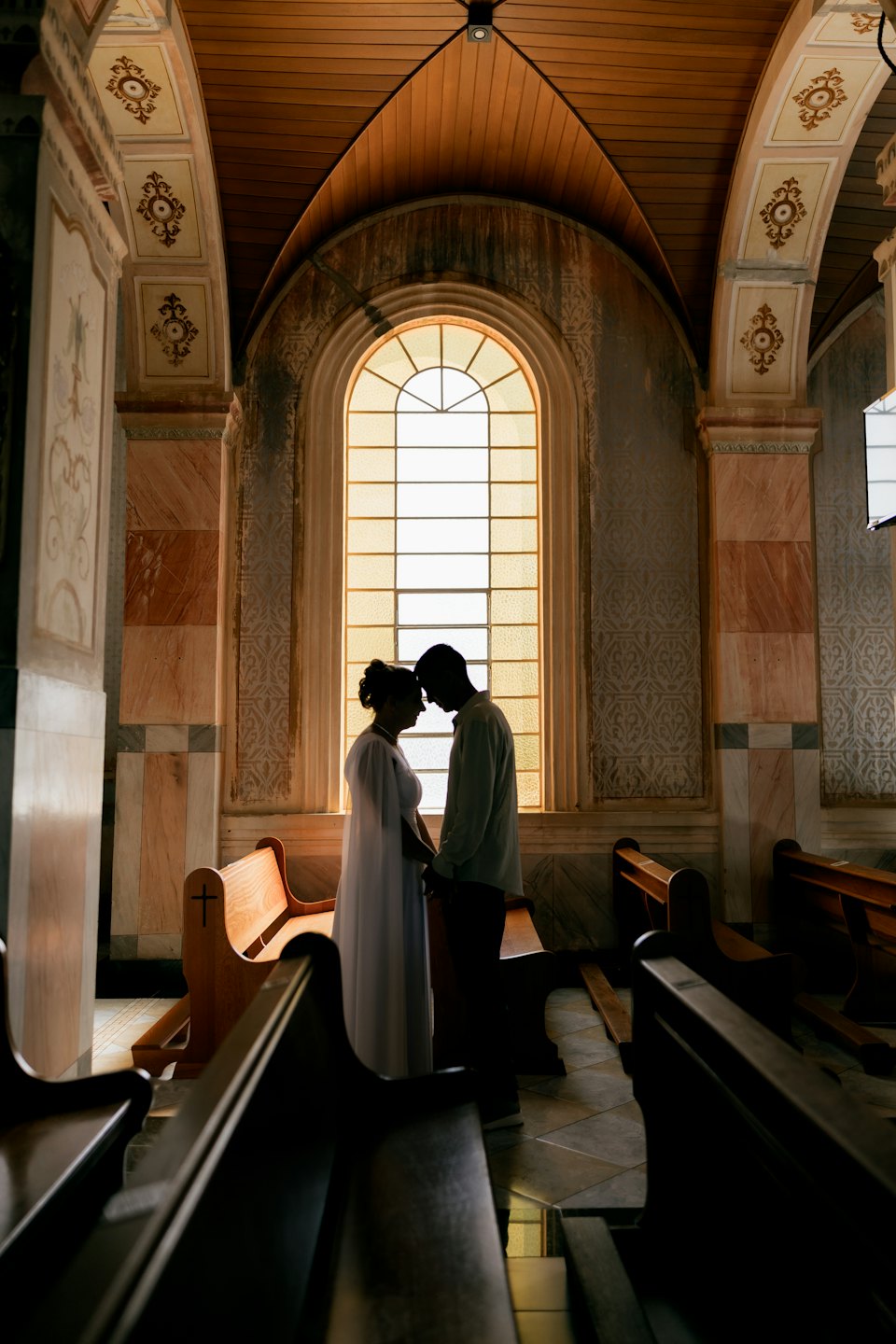 A couple holds hands and touches foreheads before a stained-glass window.