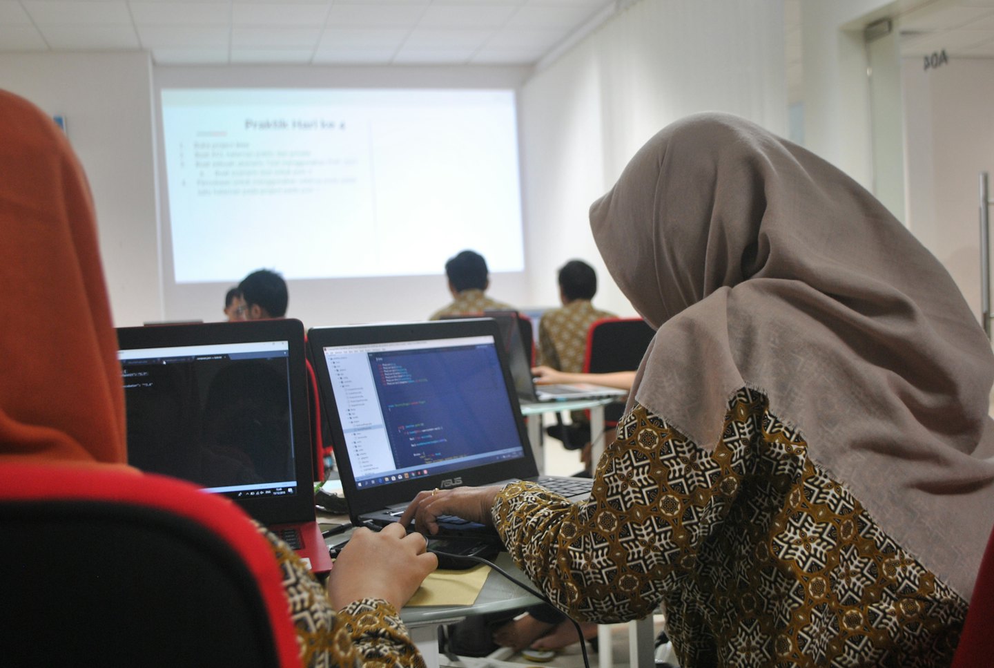 Two people type code on laptops in a bright classroom.