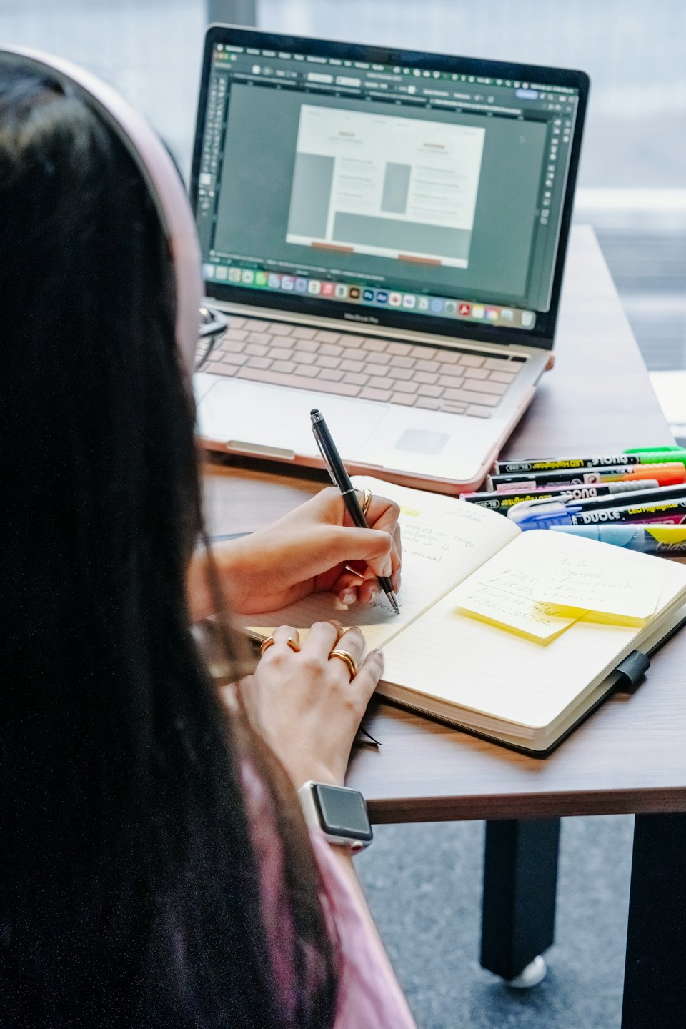 Designer sketches in notebook while working on laptop UI.